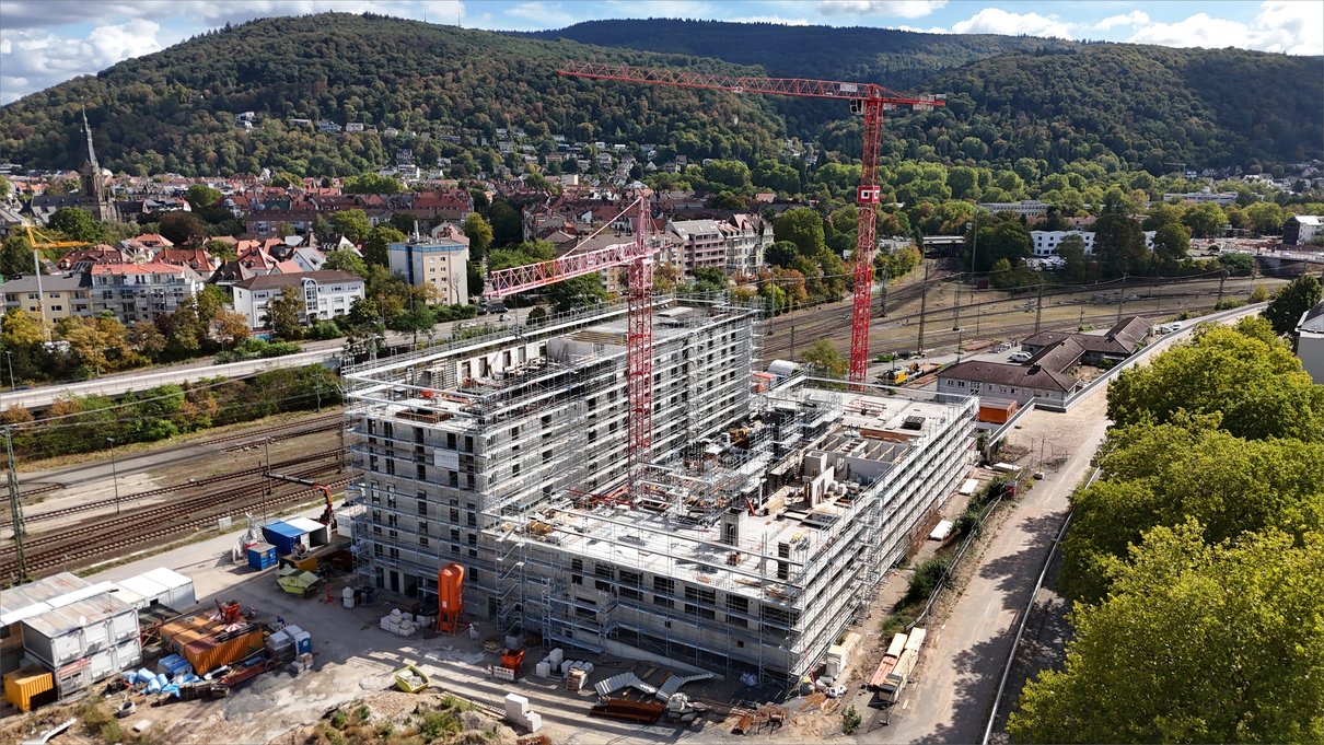 Rohbau mit umlaufendem Fassadengerüst und zwei Turmdrehkranen vor den bewaldeten Hängen über Heidelberg
