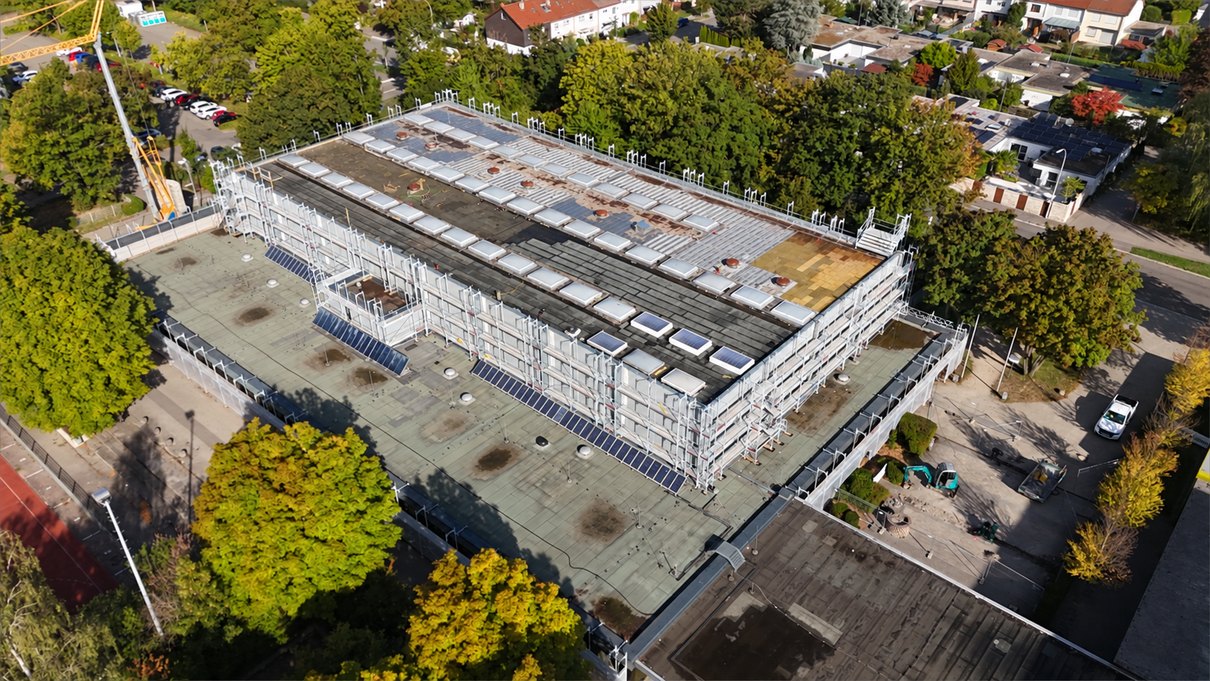 Die Elly-Schule aus größerer Höhe, das Gerüst umschließt den aufgesetzten Dachbereich, im Umfeld Sportplatz und Wohnbebauung