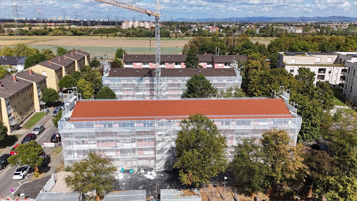 Die Wohnzeile mit fertiger Ziegeldeckung im Fassadengerüst, dahinter der zweite Bauteil und der Turmdrehkran