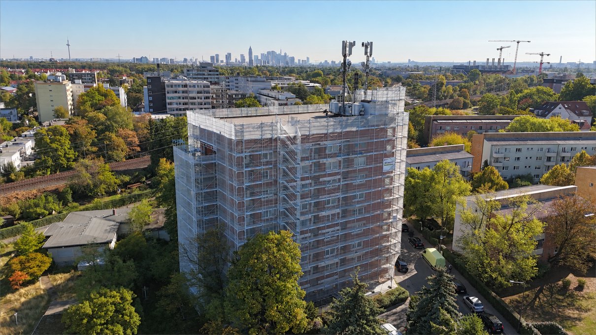 Das eingerüstete Hochhaus aus der Luft, am Horizont die Frankfurter Skyline, ringsum Wohnbebauung und Baumbestand